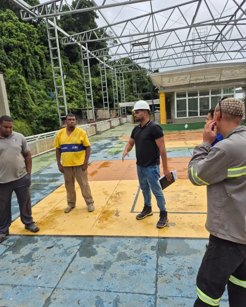 Rhuann, de capacete branco, orientando a equipe na obra sob uma estrutura metálica treliçada